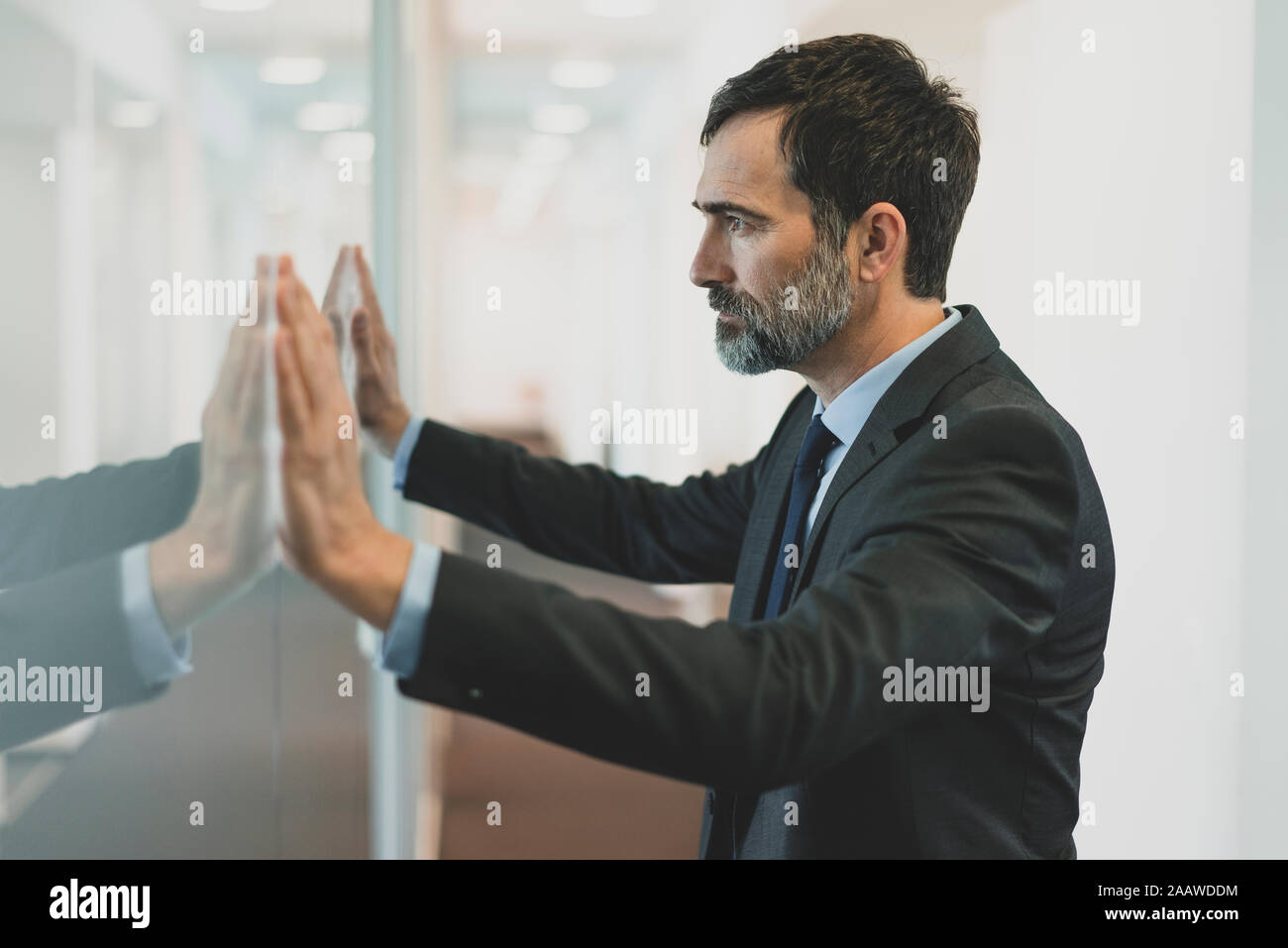 Thoughtful mature businessman leaning against glass wall in office Stock Photo