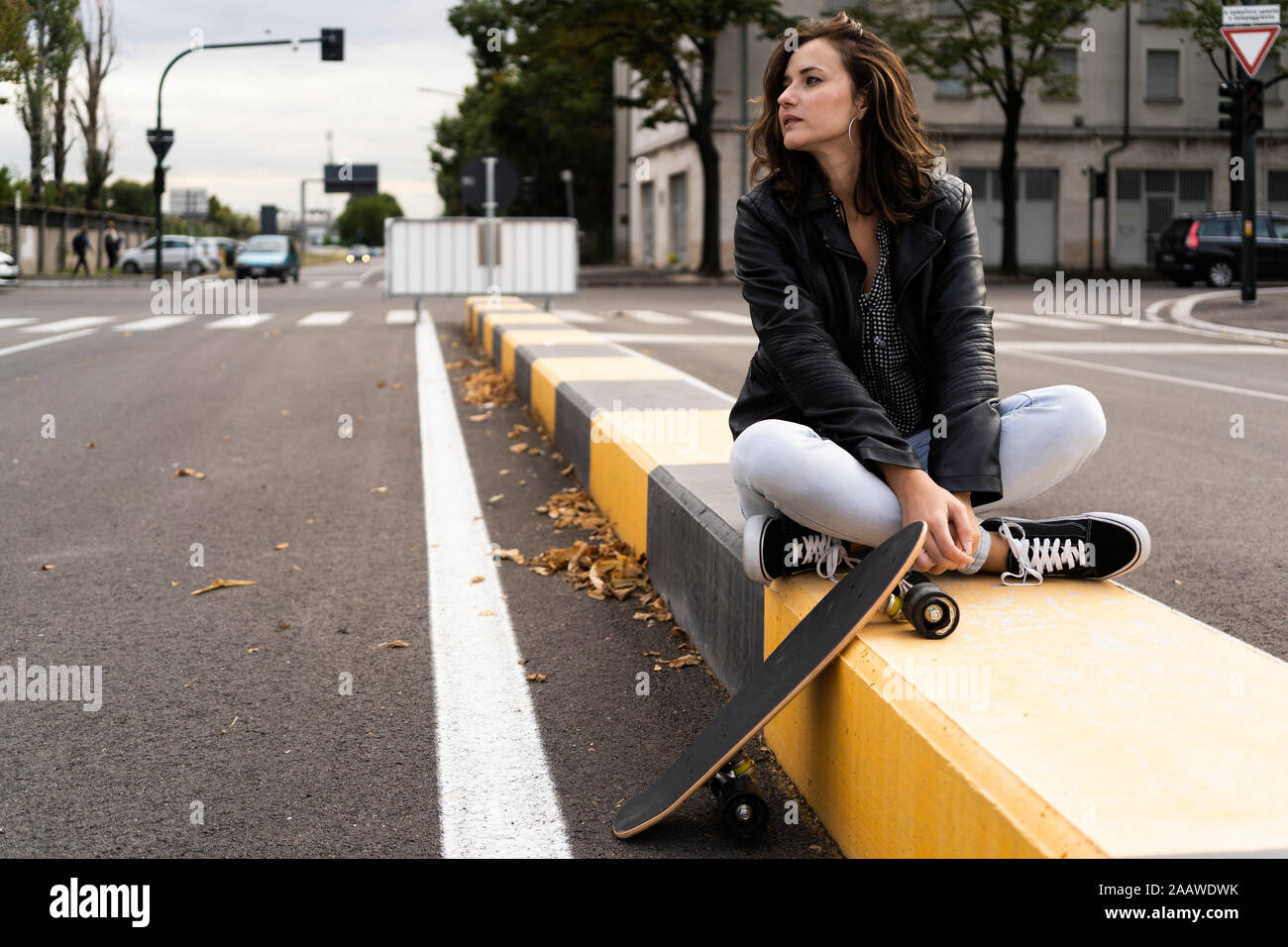 Young woman with skateboard sitting on bollard looking at distance Stock Photo