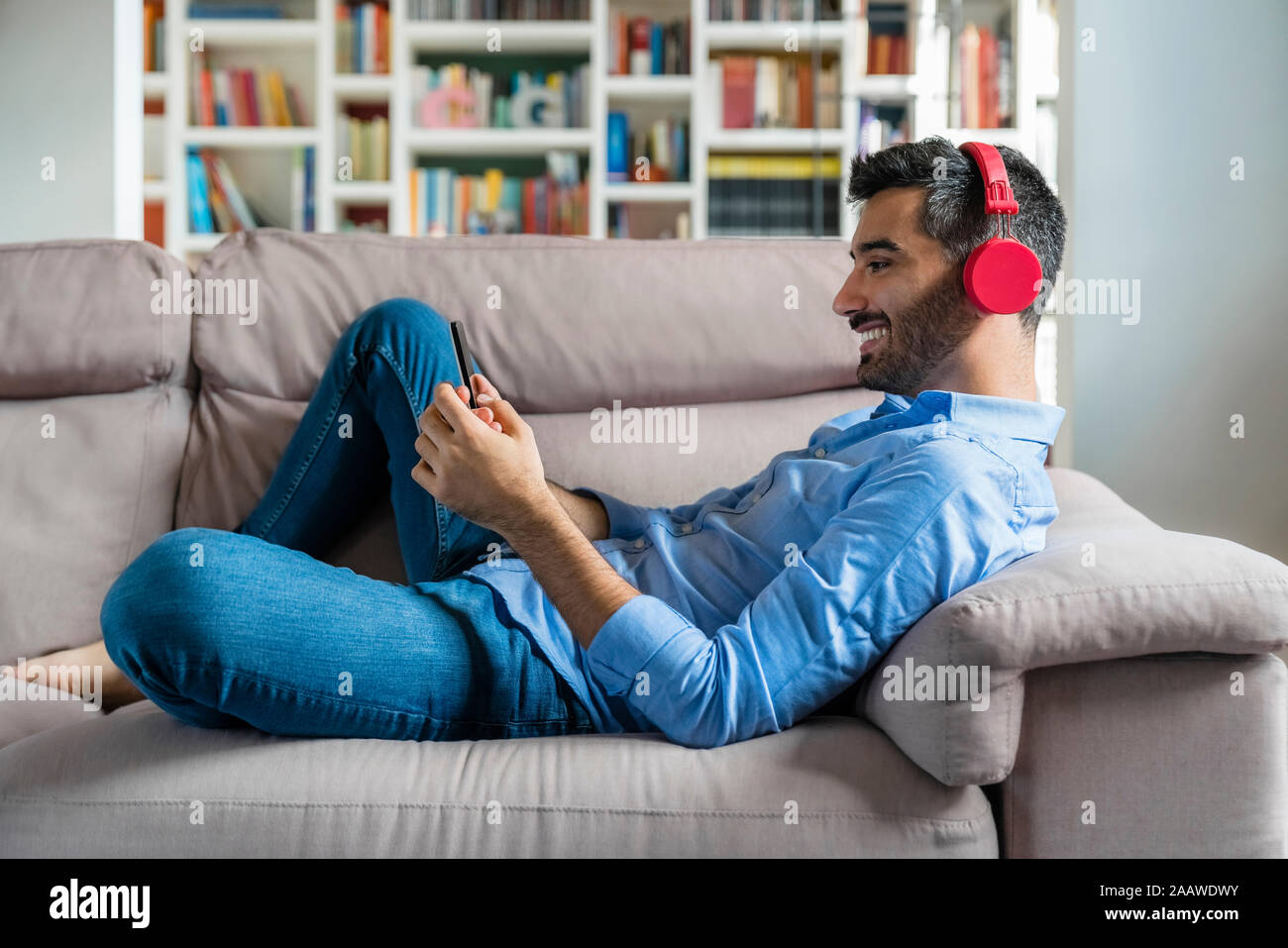 Smiling young man lying on the couch at home using smartphone and wireless headphones Stock Photo