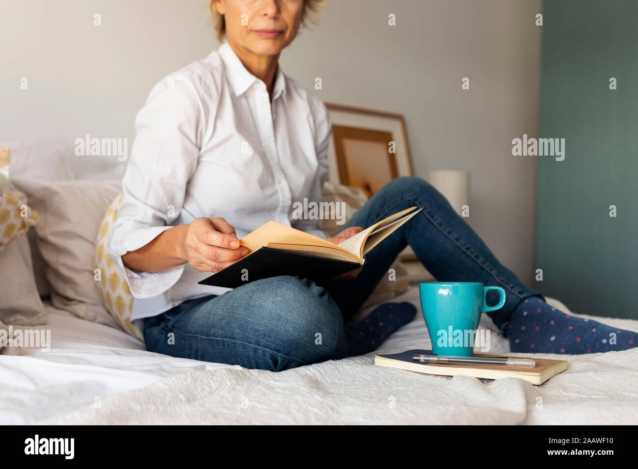 Mature woman with book and coffee cup sitting on bed at home Stock Photo
