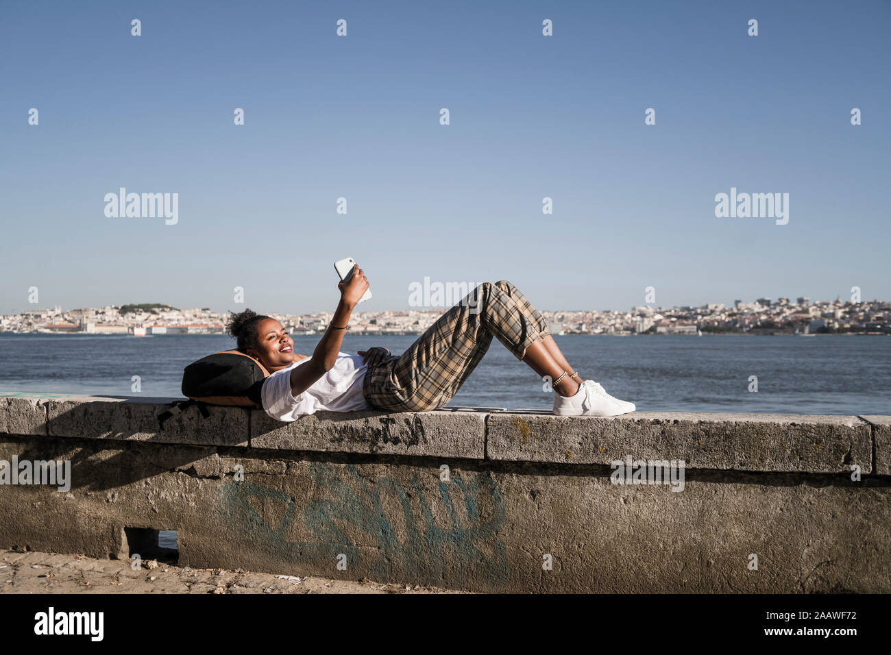 Young woman lying on a wall at the waterfront using cell phone, Lisbon, Portugal Stock Photo