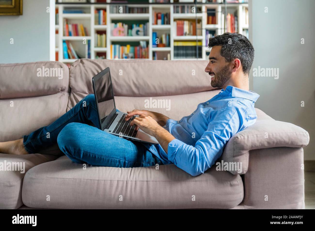 Smiling young man lying on the couch at home using laptop Stock Photo