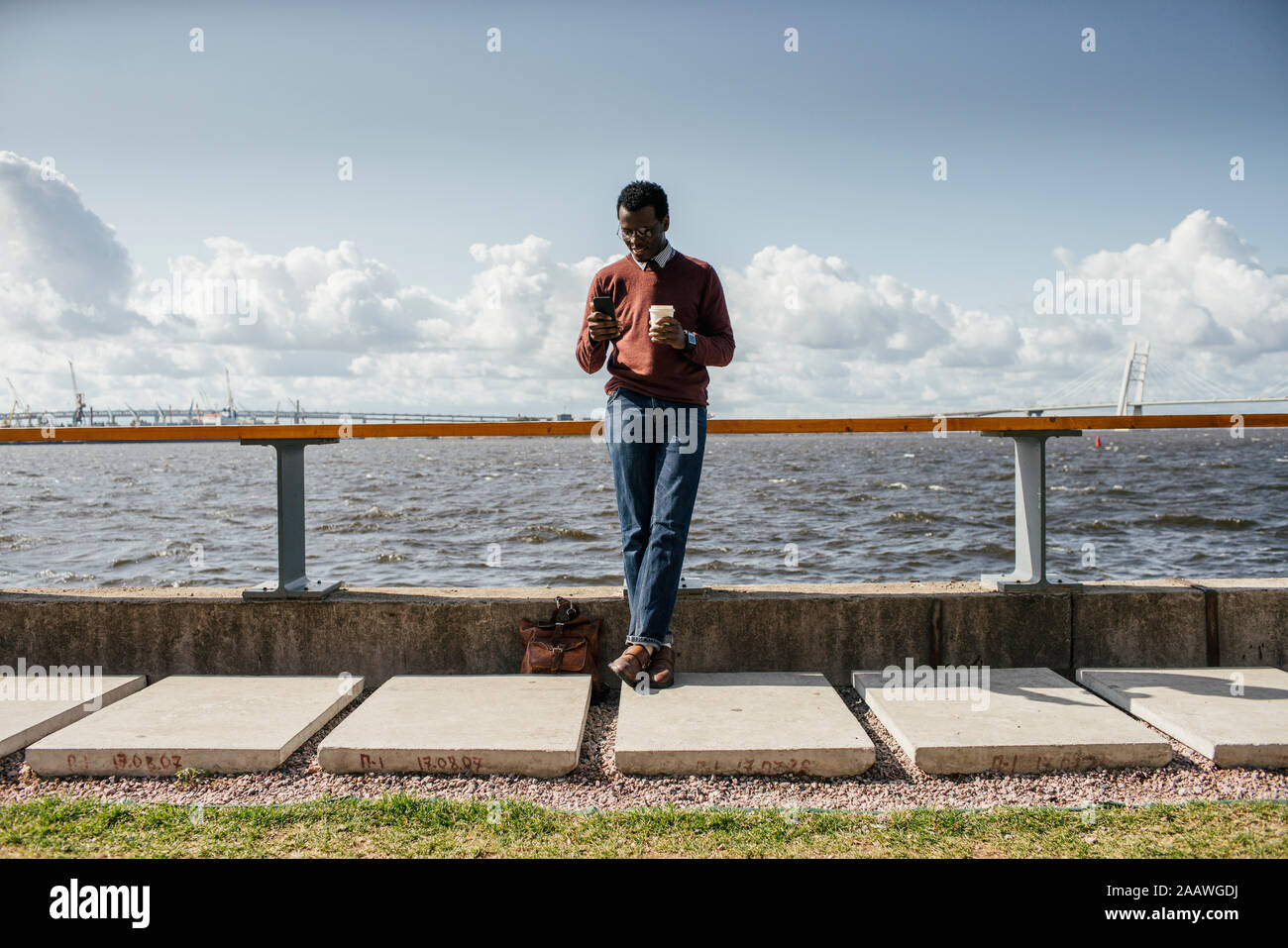 Young man standing at the sea, holding coffee, using smartphone Stock Photo