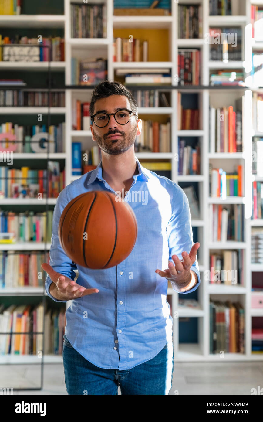 Portrait of young man with basketball standing in front of bookshelves at home Stock Photo