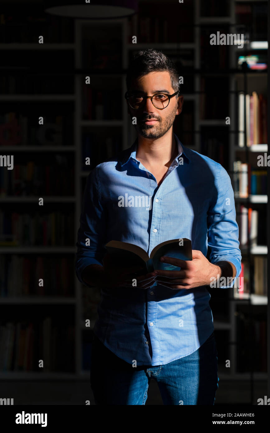 Portrait of young man with book standing in front of bookshelves at home Stock Photo