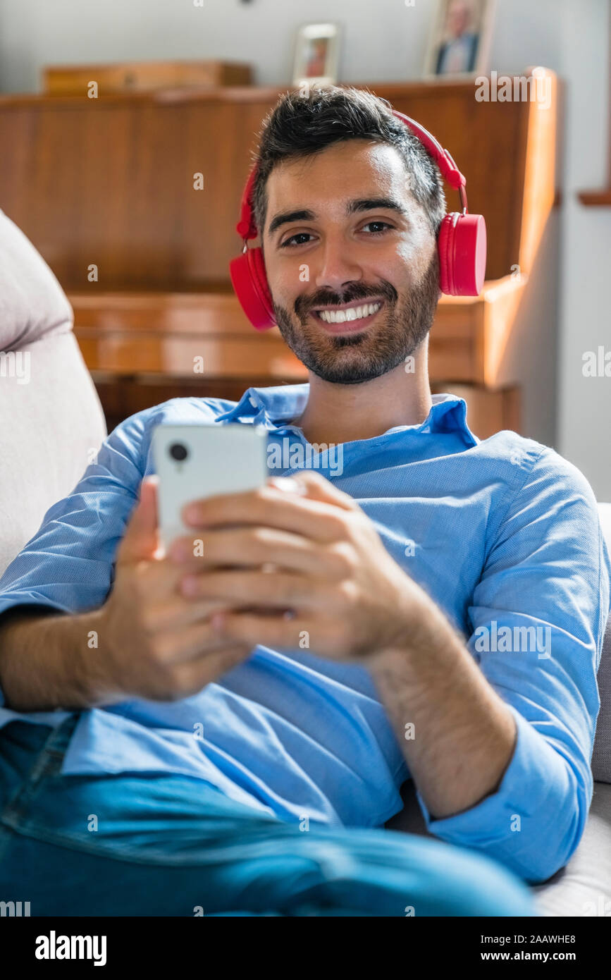 Portrait of smiling young man lying on the couch at home using smartphone and wireless headphones Stock Photo