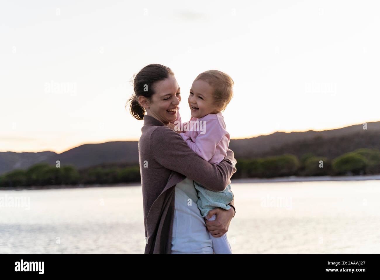 Happy mother carrying her daughter on a jetty at sunset Stock Photo