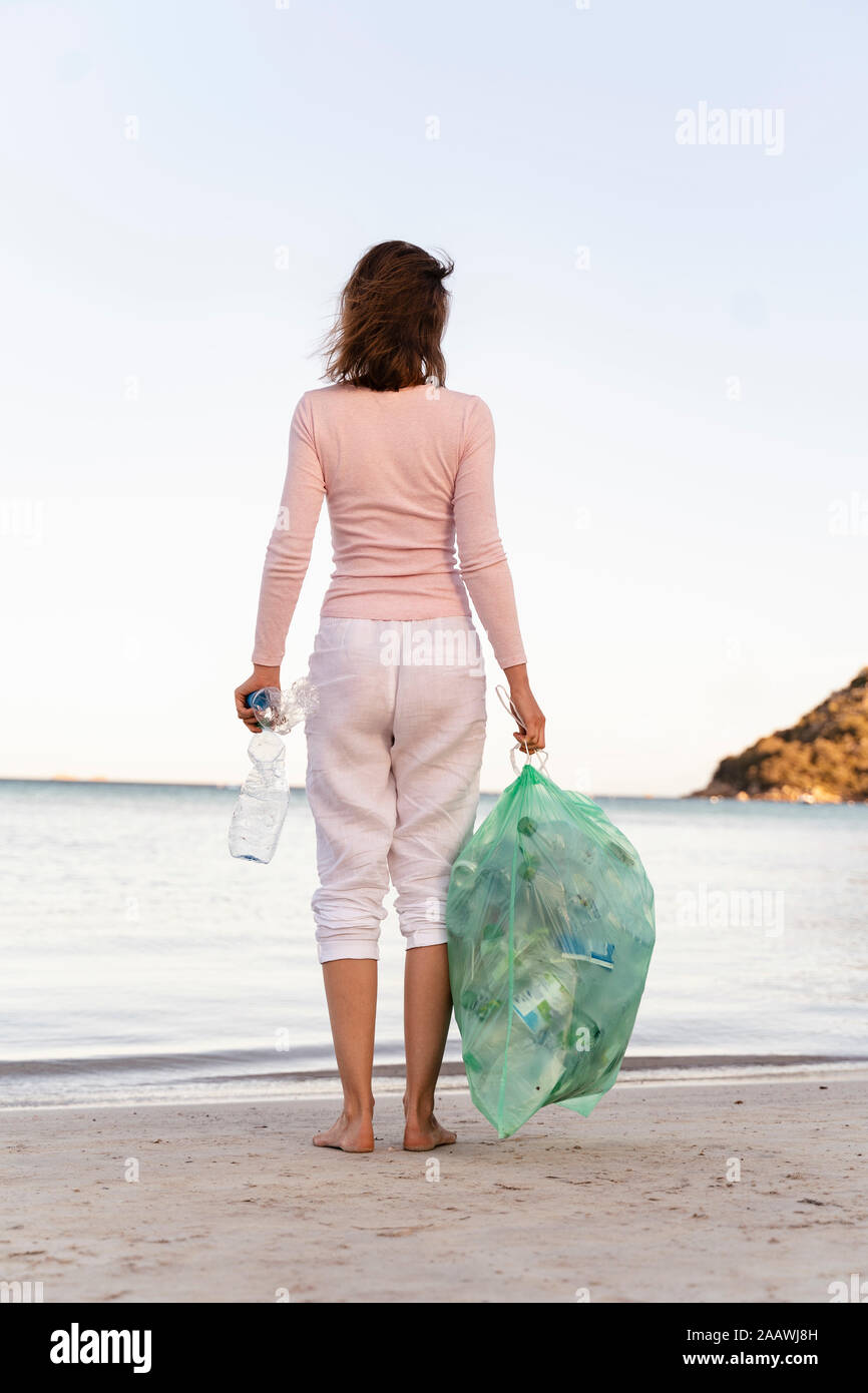 Back view of woman standing on the beach with bin bag of collected empty plastic bottles Stock Photo