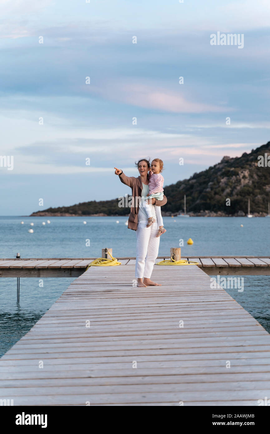 Happy mother carrying daughter on a jetty at sunset Stock Photo