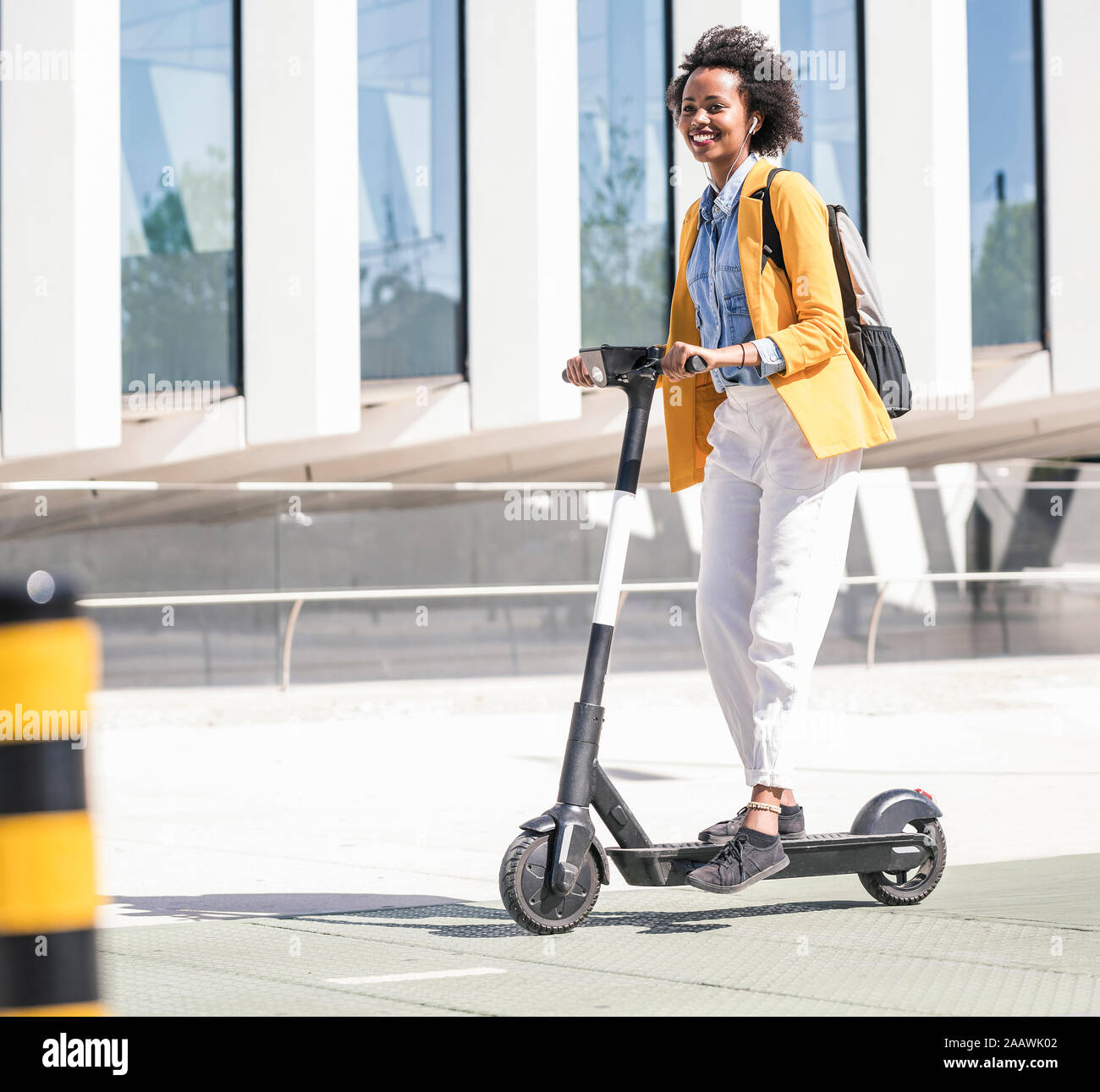 Happy young woman with earphones riding e-scooter in the city Stock Photo