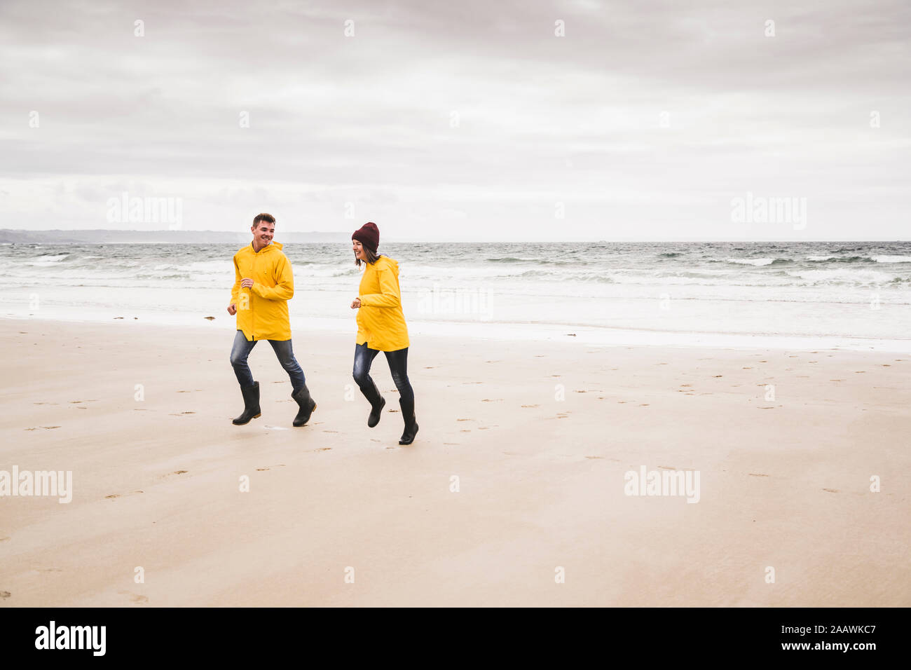 Young woman wearing yellow rain jackets and running at the beach, Bretagne, France Stock Photo