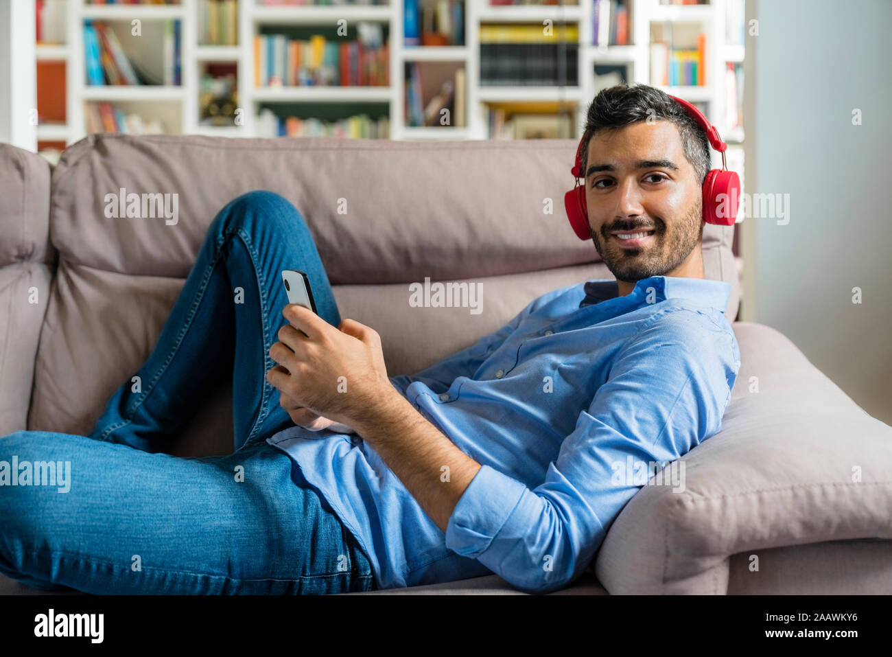 Portrait of smiling young man lying on the couch at home using smartphone and wireless headphones Stock Photo