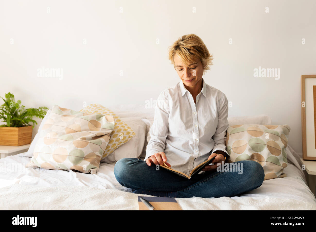 Mature woman with book sitting on bed at home Stock Photo