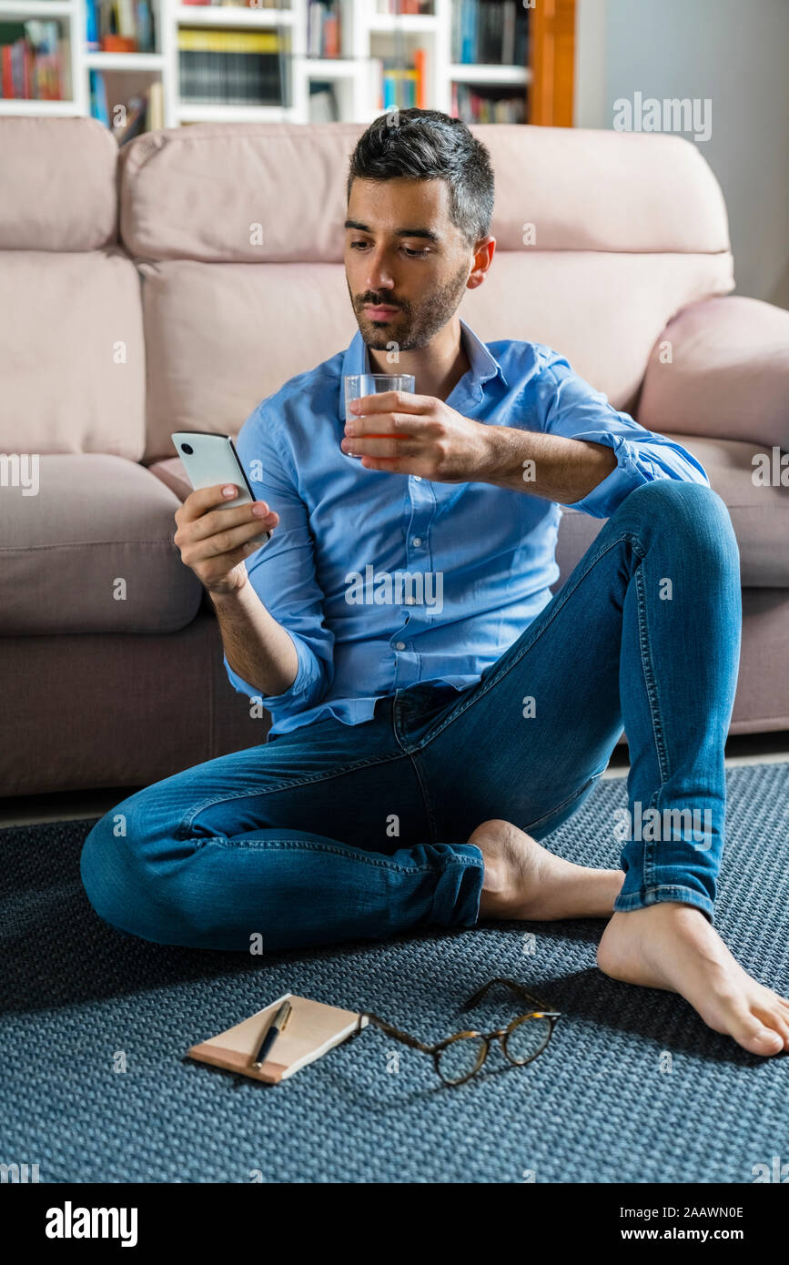 Portrait of young man sitting barefoot on the floor of living room at home looking at smartphone Stock Photo