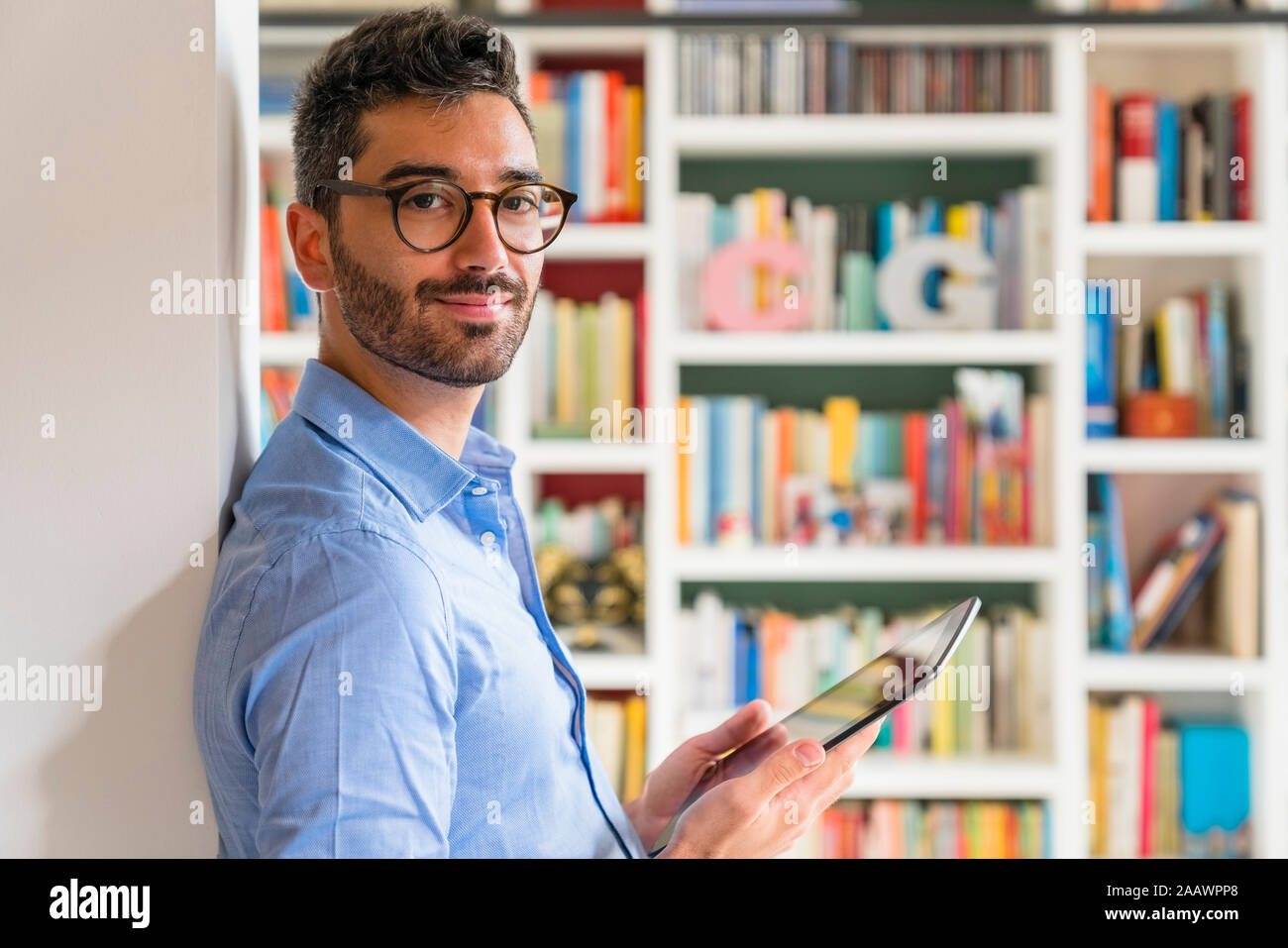 Portrait of smiling young man standing in front of bookshelves at home with digital tablet Stock Photo