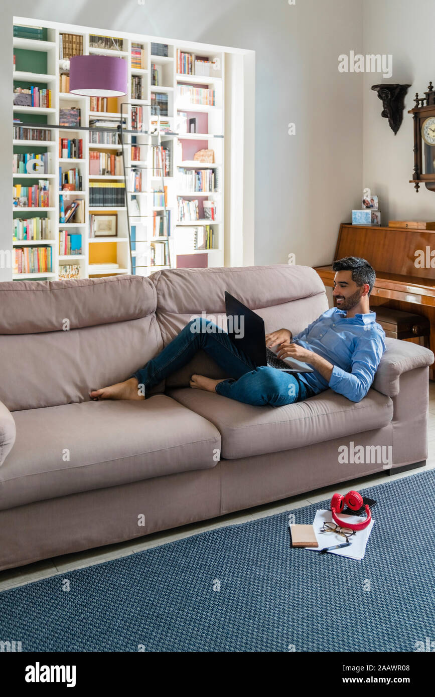 Smiling young man lying on the couch at home using laptop Stock Photo