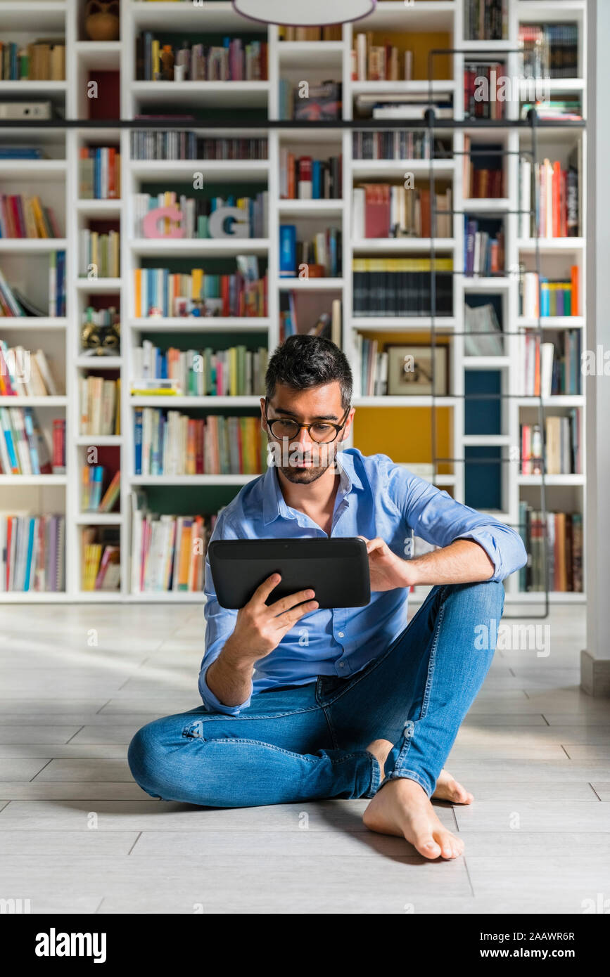Portrait of barefoot young man sitting in front of bookshelves on the floor using digital tablet Stock Photo