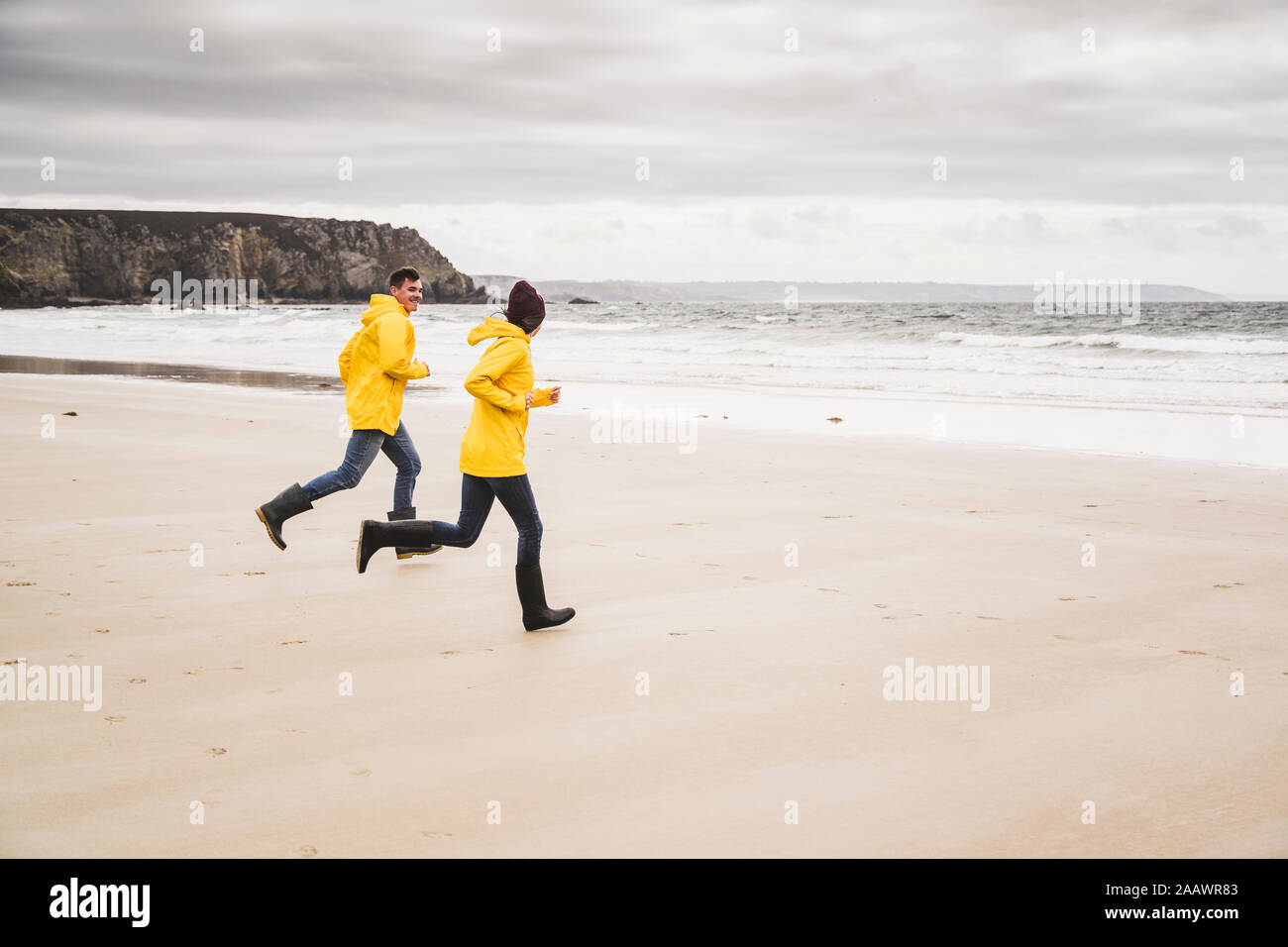 Young woman wearing yellow rain jackets and running at the beach, Bretagne, France Stock Photo