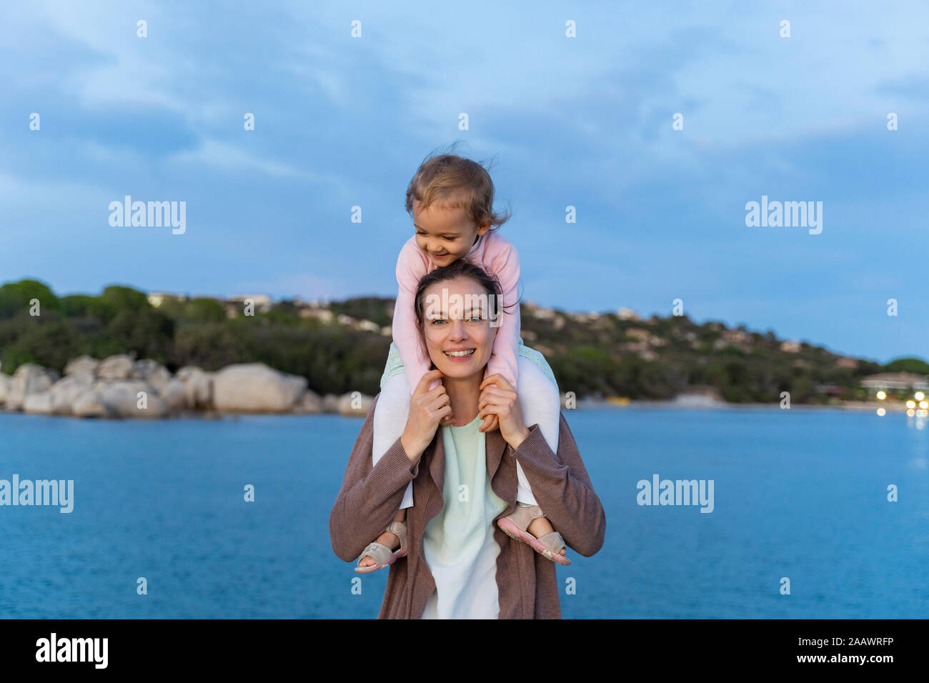 Portrait of mother carrying her daughter on shoulders at the seafront at sunset Stock Photo