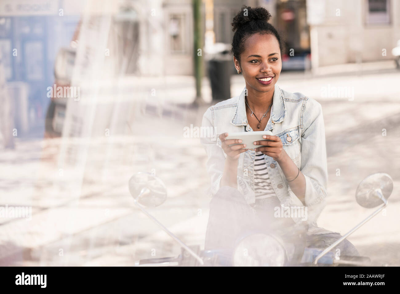 Smiling young woman with motor scooter and mobile phone in the city, Lisbon, Portugal Stock Photo