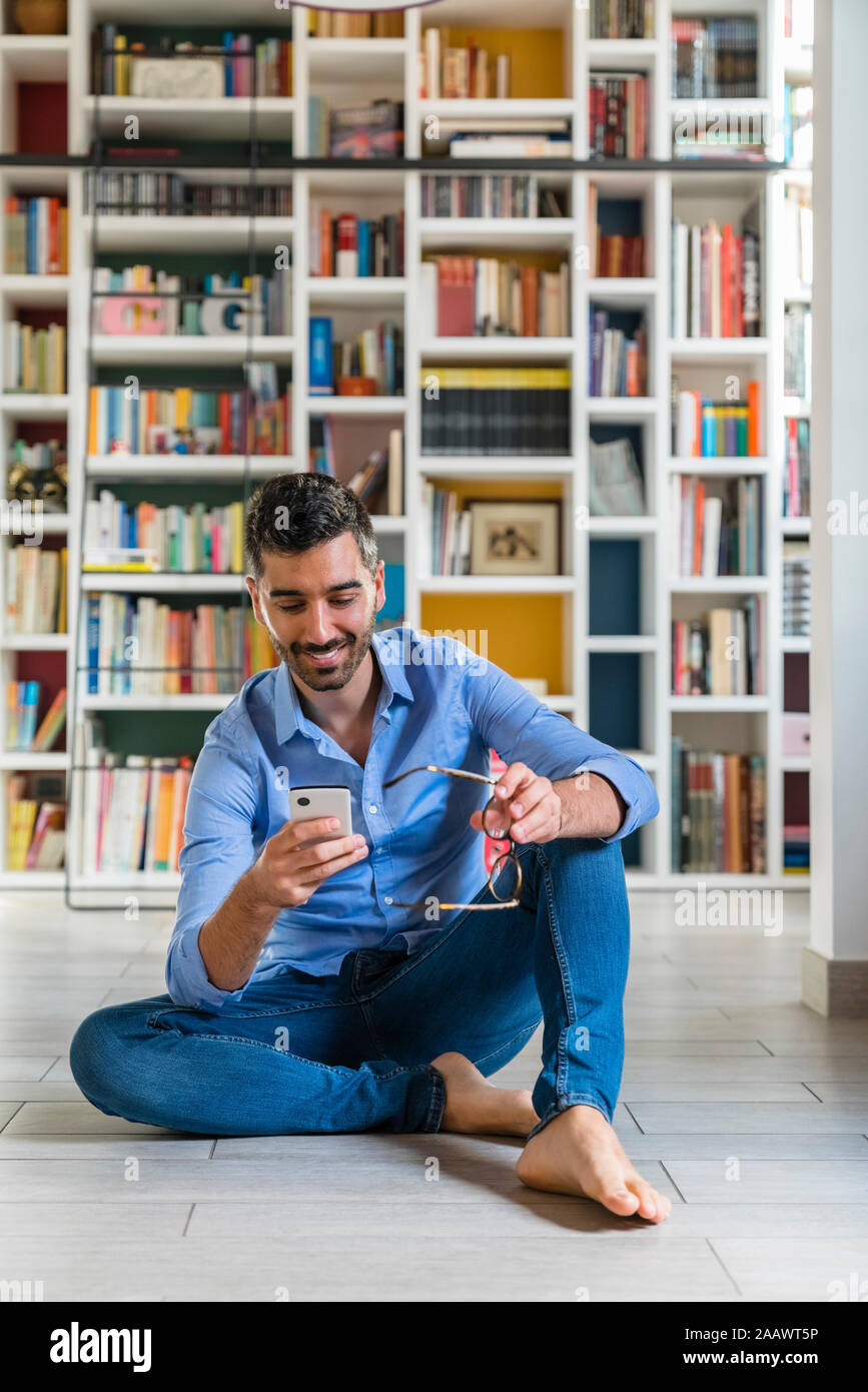 Portrait of smiling young man sitting in front of bookshelves on the floor at home looking at smartphone Stock Photo