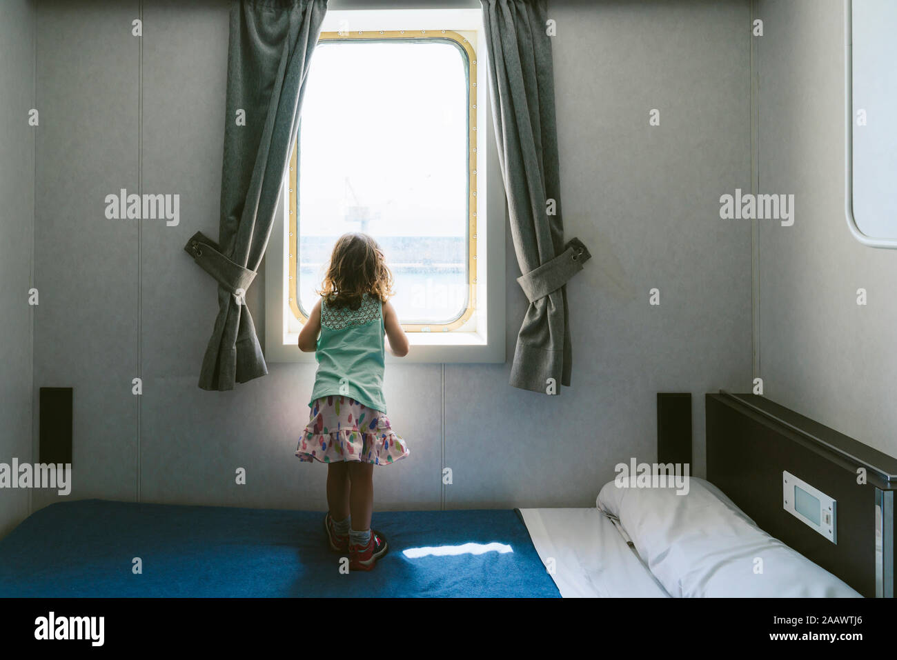 Back view of little girl standing on bed looking to the sea from window of ship's cabin Stock Photo