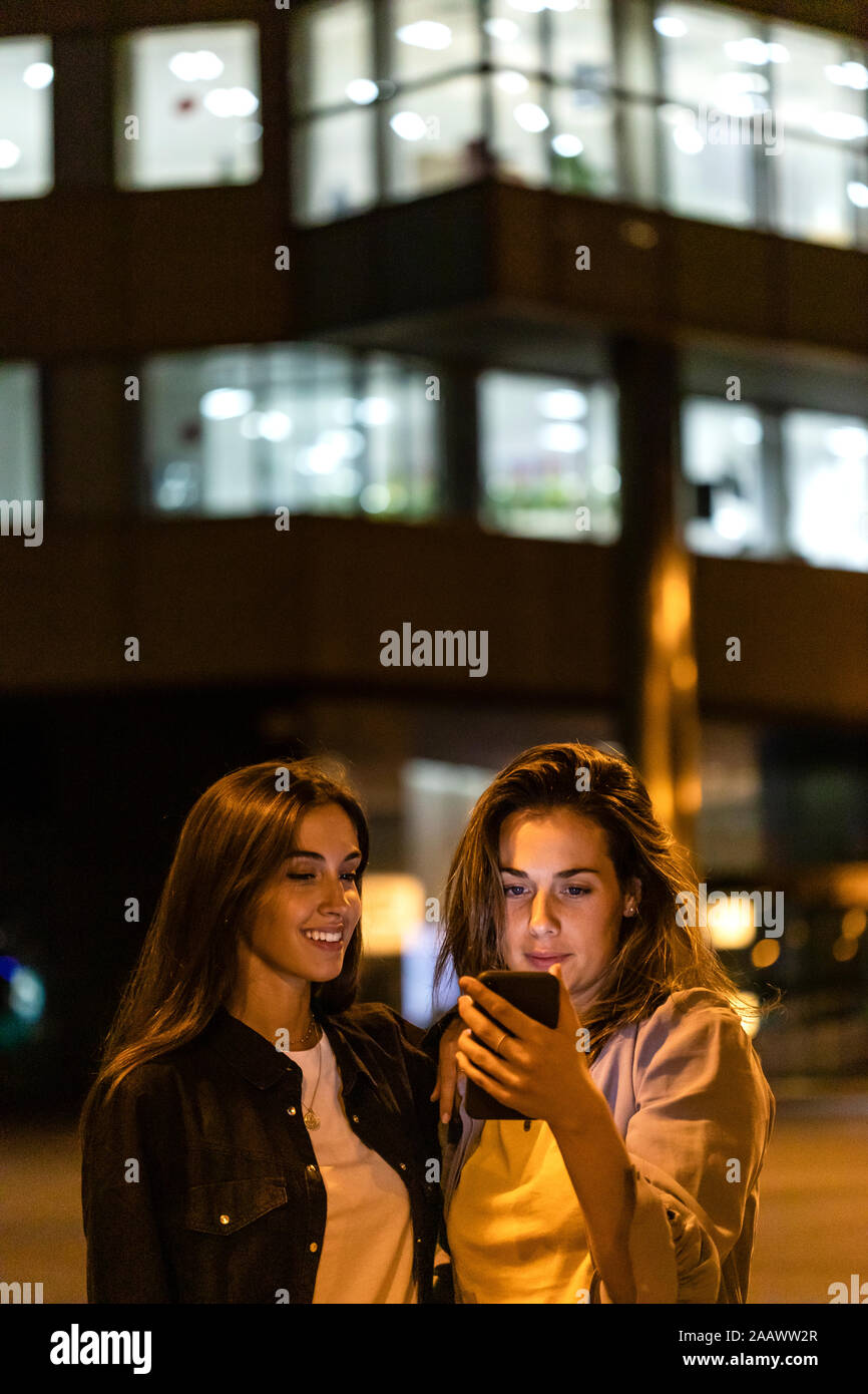 Two friends using the smartphone at night, with city lights in the background Stock Photo