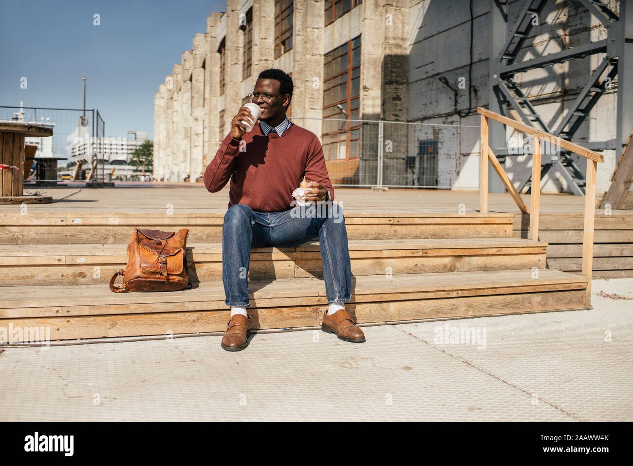 Young man sitting on stairs in the city, eating hamburger, drinking coffee Stock Photo