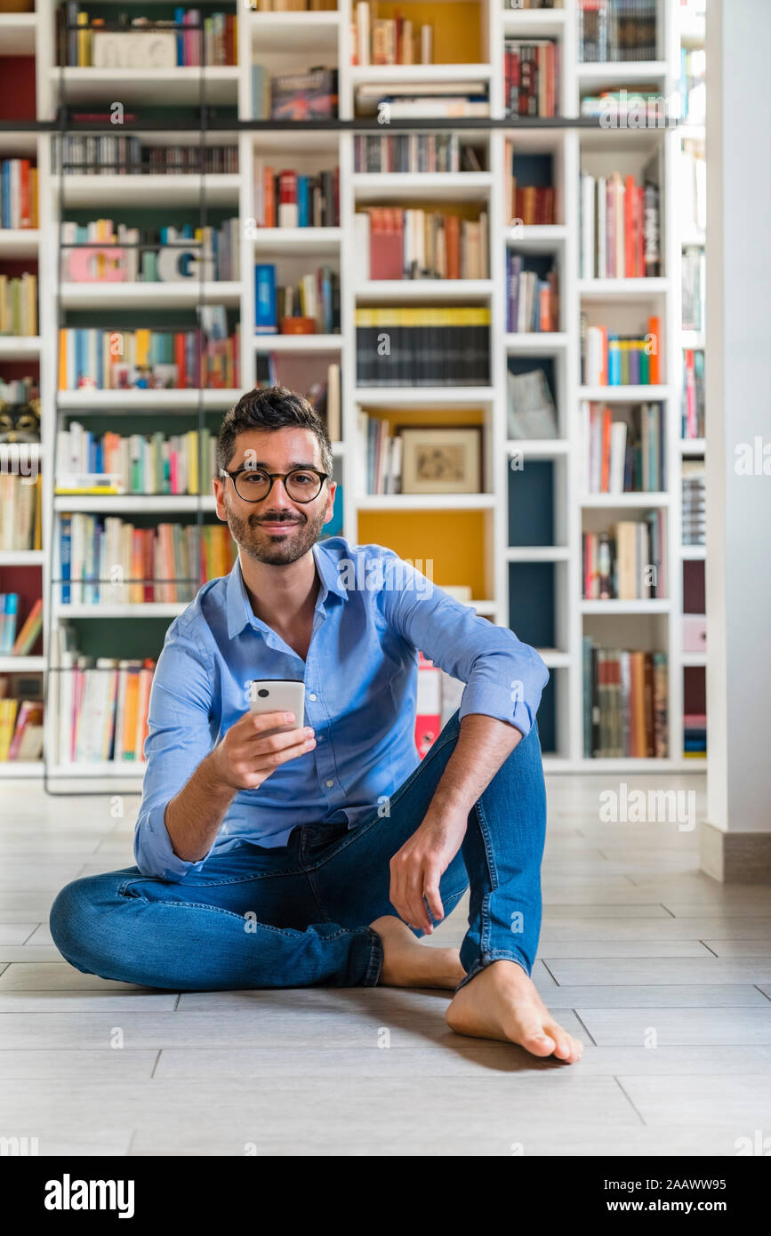 Portrait of smiling young man sitting with smartphone in front of bookshelves on the floor at home Stock Photo