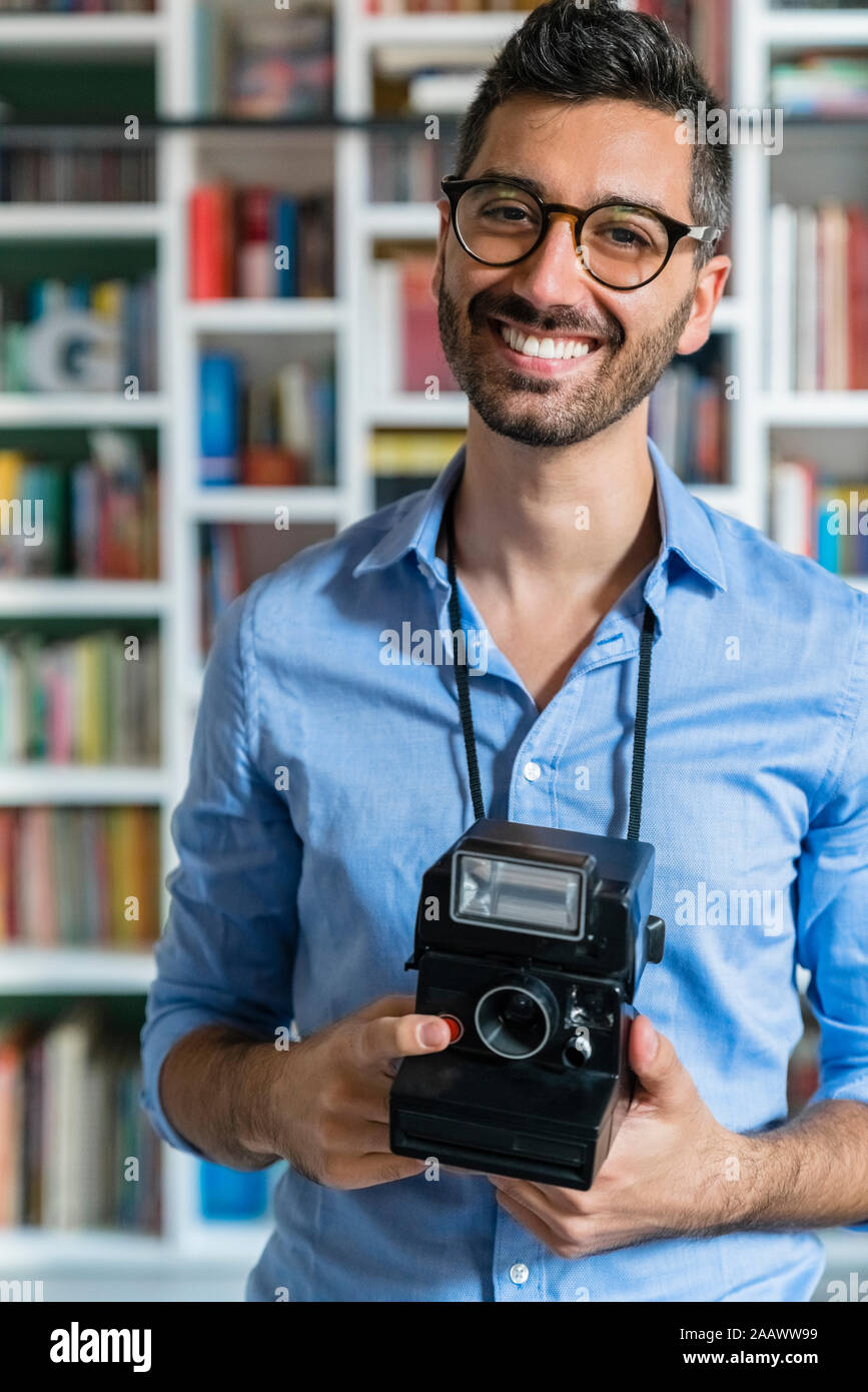 Portrait of happy young man with instant camera standing in front of bookshelves Stock Photo