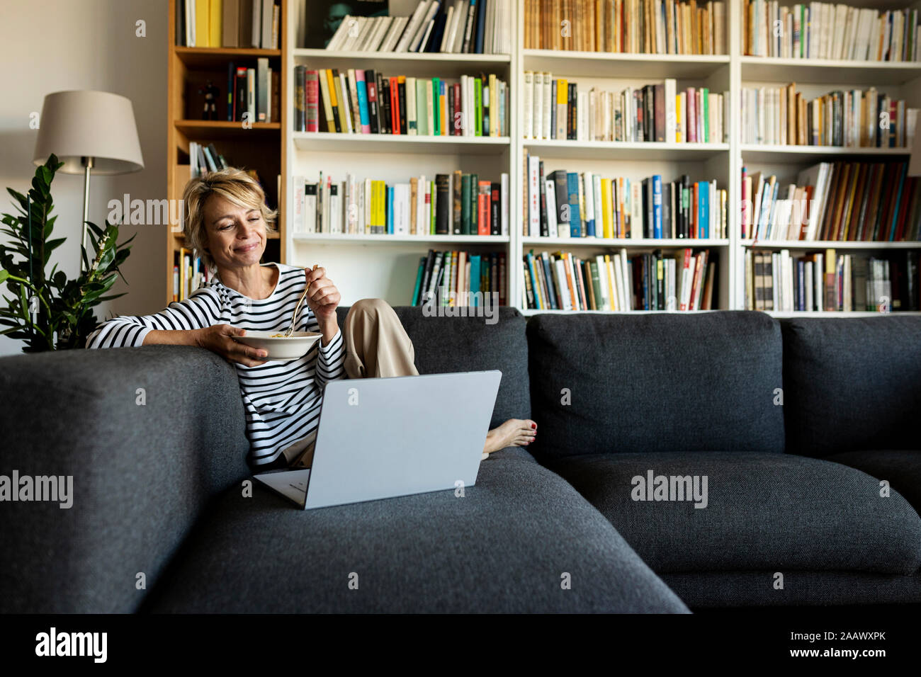 Mature woman using laptop and having lunch on couch at home Stock Photo