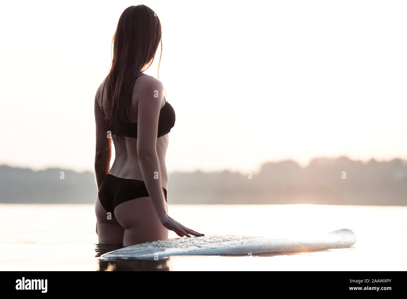 Back view of young woman with surfboard standing at shore of Lake Starnberg watching sunset, Germany Stock Photo