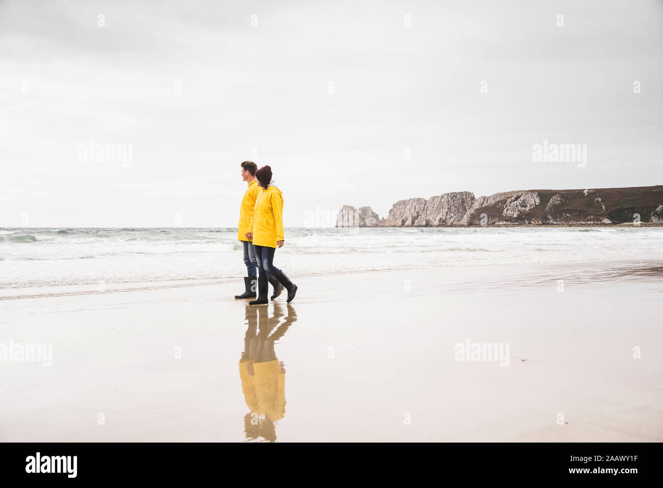 Young woman wearing yellow rain jackets and walking along the beach, Bretagne, France Stock Photo