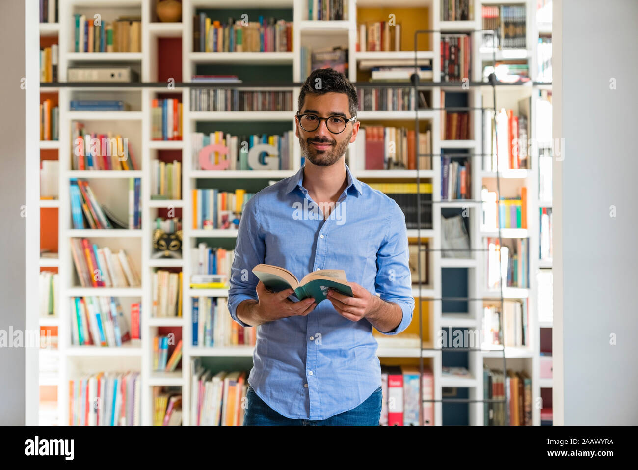 Portrait of smiling young man with book standing in front of bookshelves at home Stock Photo