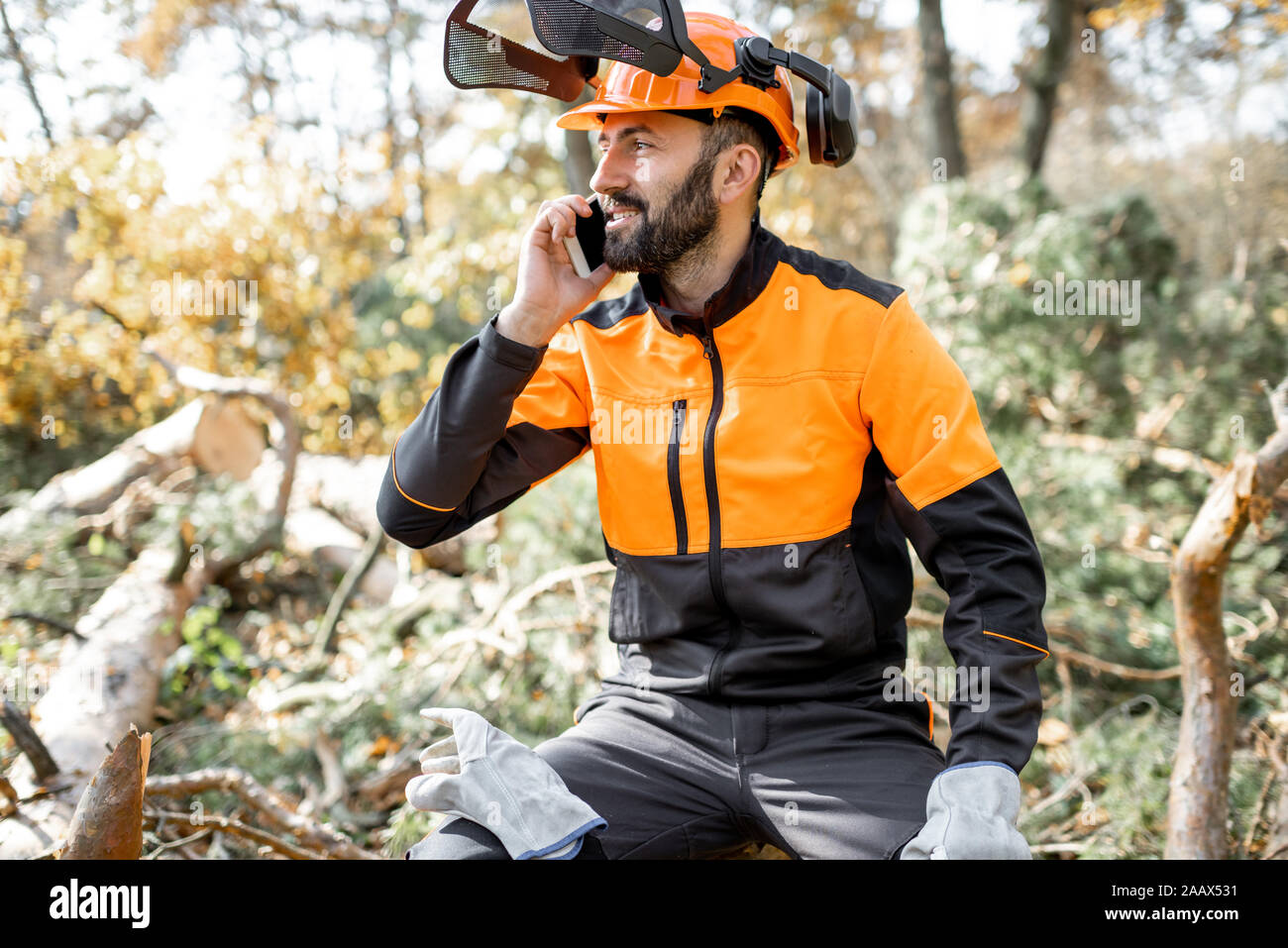 Professional lumberman in protective workwear talking on phone, while sitting on the felled tree, resting after the hard work in the forest Stock Photo
