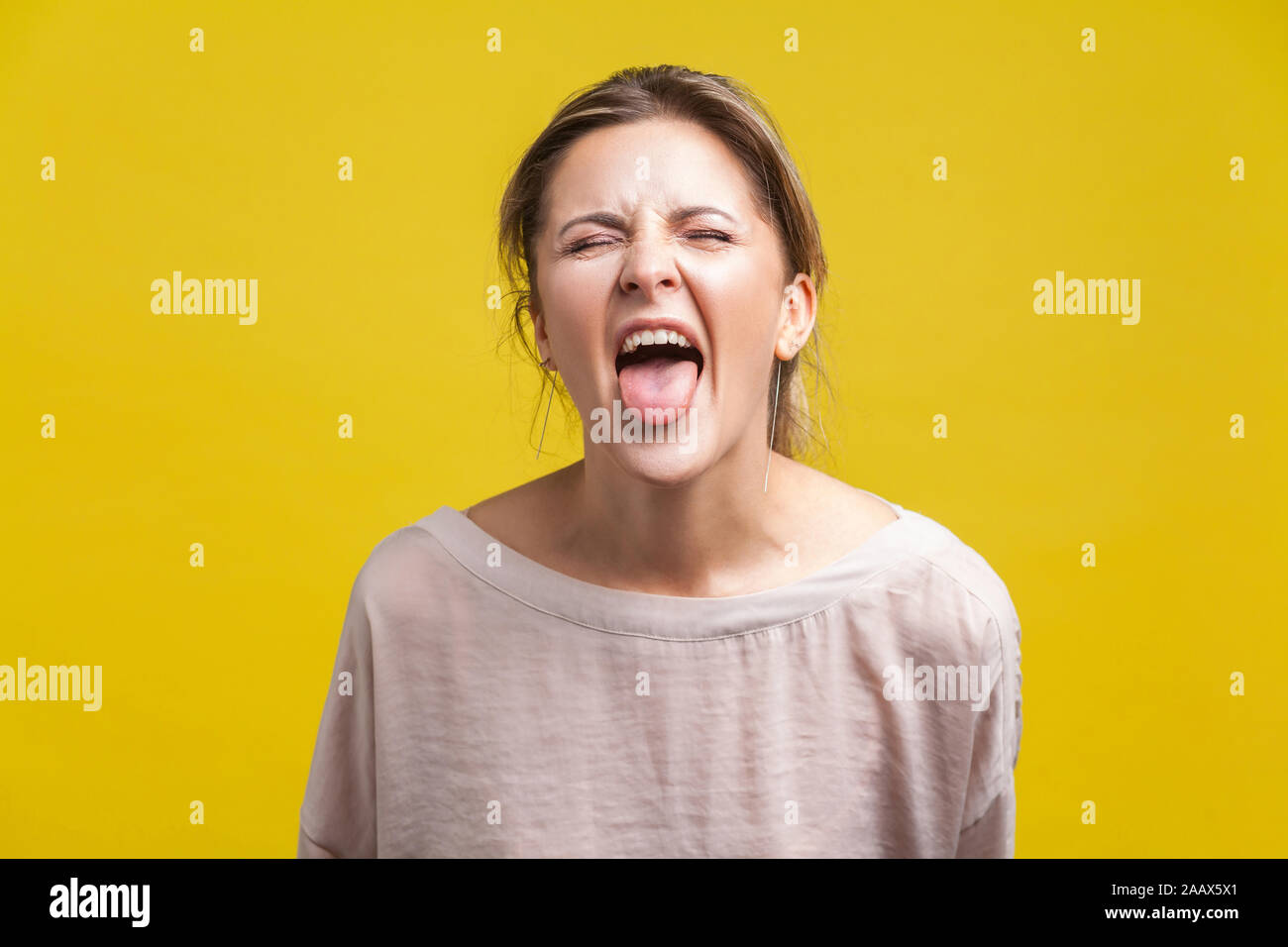 Portrait of young naughty woman with blonde hair in casual beige blouse standing with closed eyes and tongue out, teasing grimace, facial expression. Stock Photo