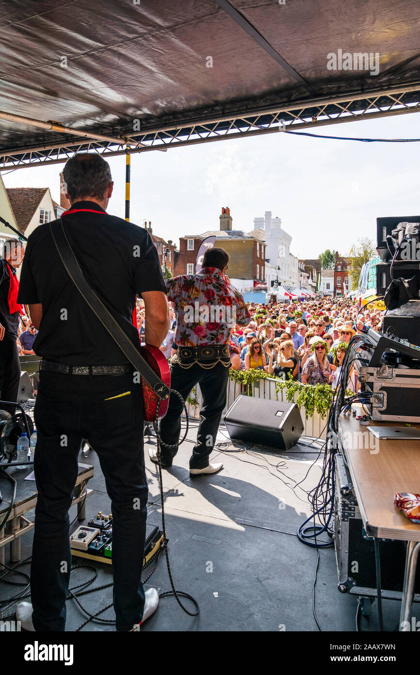 Faversham Hop Festival. View from back of stage of the 'Taking Care of Elvis' rock band on stage in front of a packed crowd during a sunny day. Stock Photo