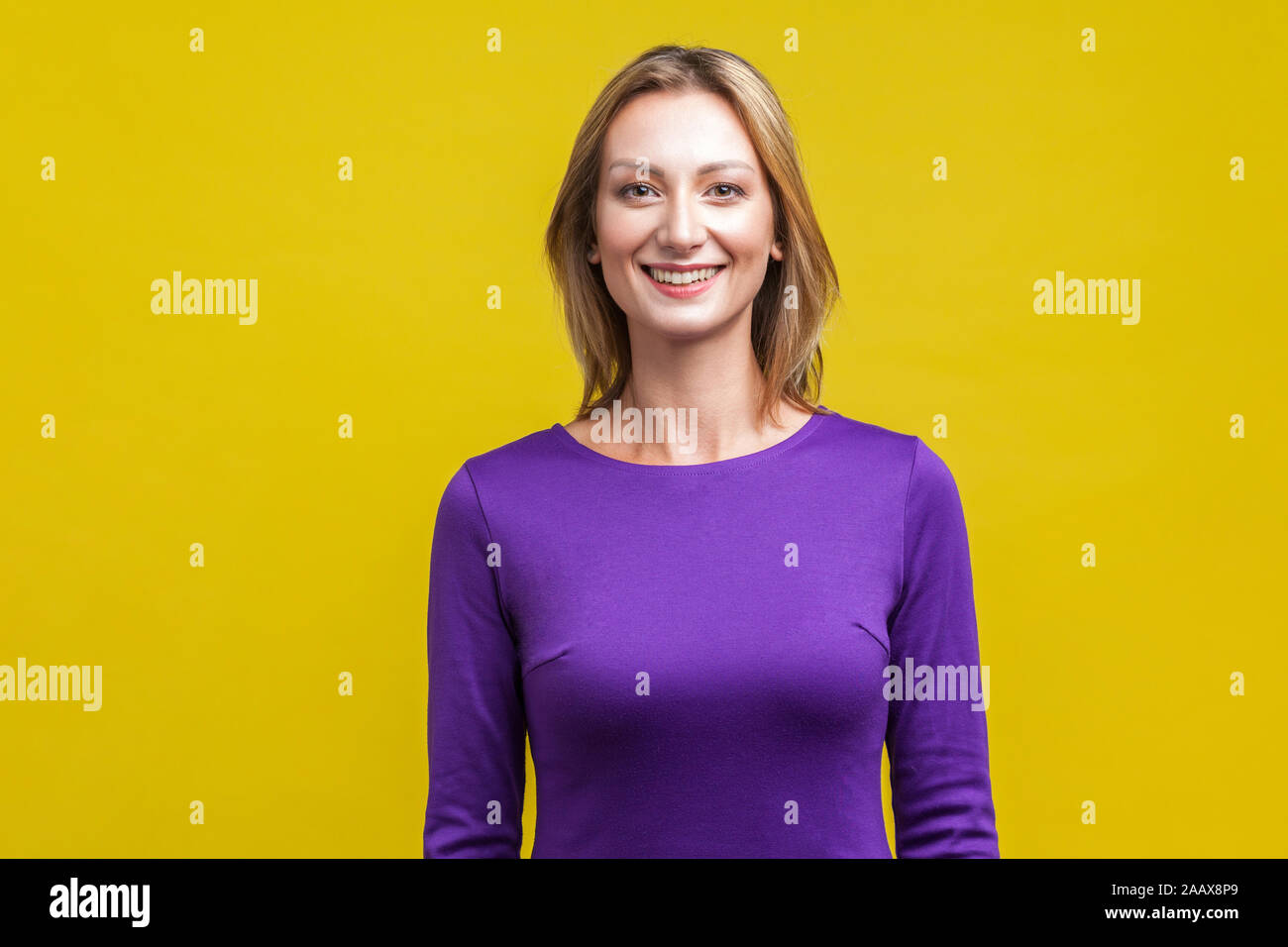 Portrait of positive elegant woman in tight purple dress standing, looking at camera with cute engaging toothy smile, beautiful with clean shiny skin. Stock Photo