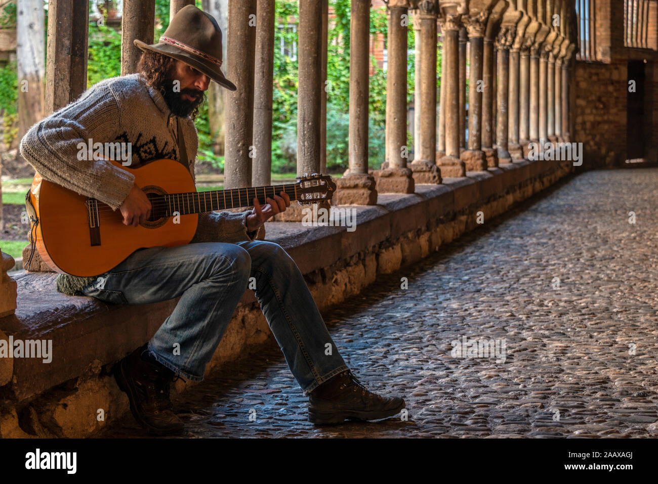 A bearded guitarist with felt hat, playing a Spanish or classical guitar, in the medieval cloisters of St Salvy church, Albi, southern France. Stock Photo