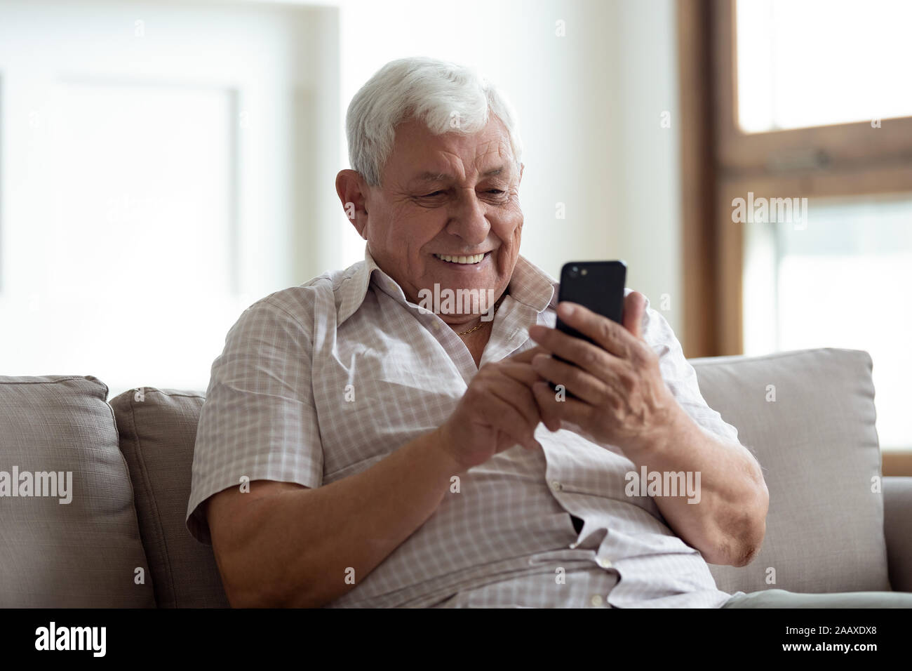 Happy older man sitting on sofa at home, using smartphone. Stock Photo
