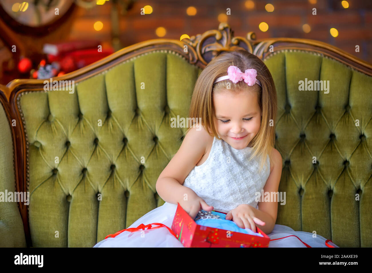 Cute little girl in a dress sitting on a sofa with a gift box in her ...