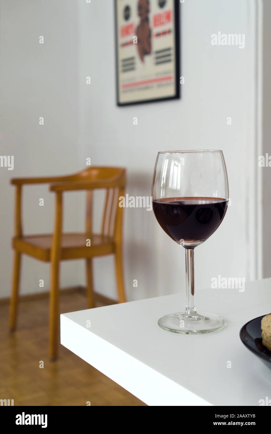 Glass of red wine on white table, blurred background with wooden chair in the room corner, domestic interior Stock Photo