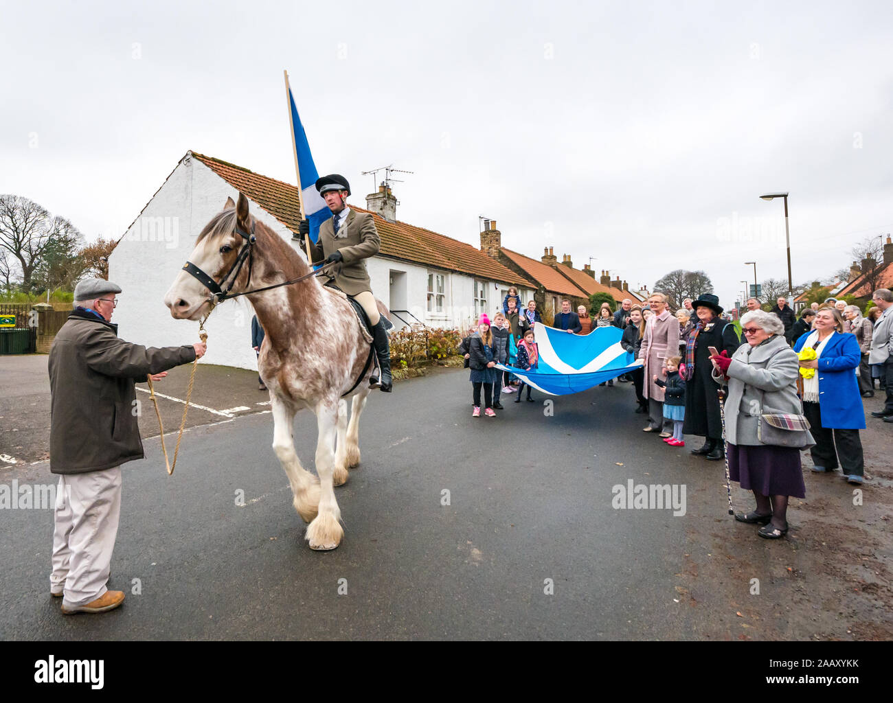 Athelstaneford, East Lothian, Scotland, UK. 24 November 2019. Saltire Festival: The first day of the annual festival to mark St Andrew’s Day takes place in the birthplace of the Scottish national flag where the saltire appeared in the sky as a good omen when the Picts & Scots defeated the Saxons led by Athelstan. Pictured: Craig Donnelly on a Strawberry Roan Clydesdale mare with groom/owner Arthur Greenan with villagers including children parade along the main street carrying a giant saltire flag Stock Photo