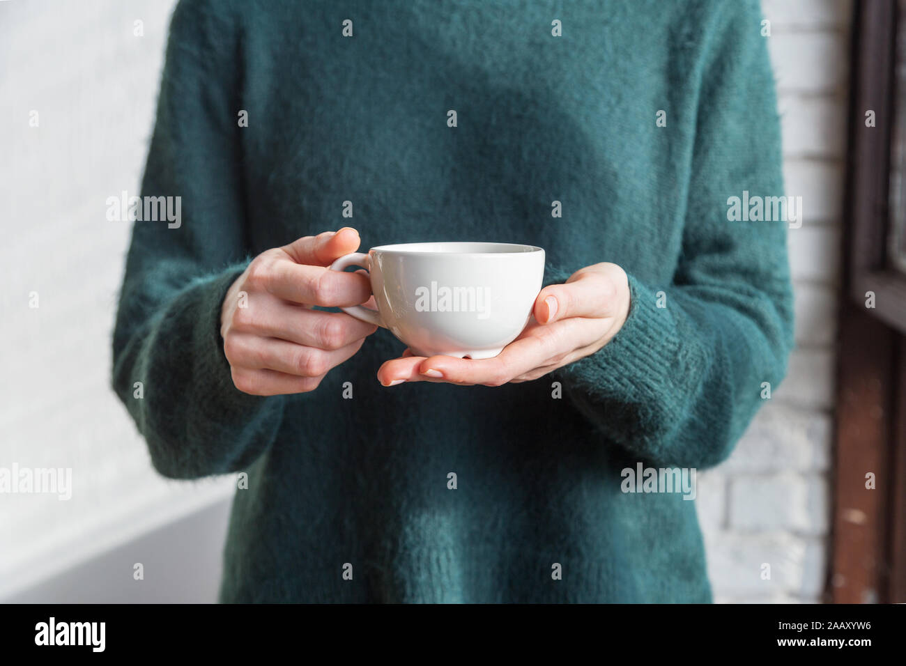 Tea cup in women's hand close up. Girl in green sweater with white cup in hands with tea or coffee Stock Photo