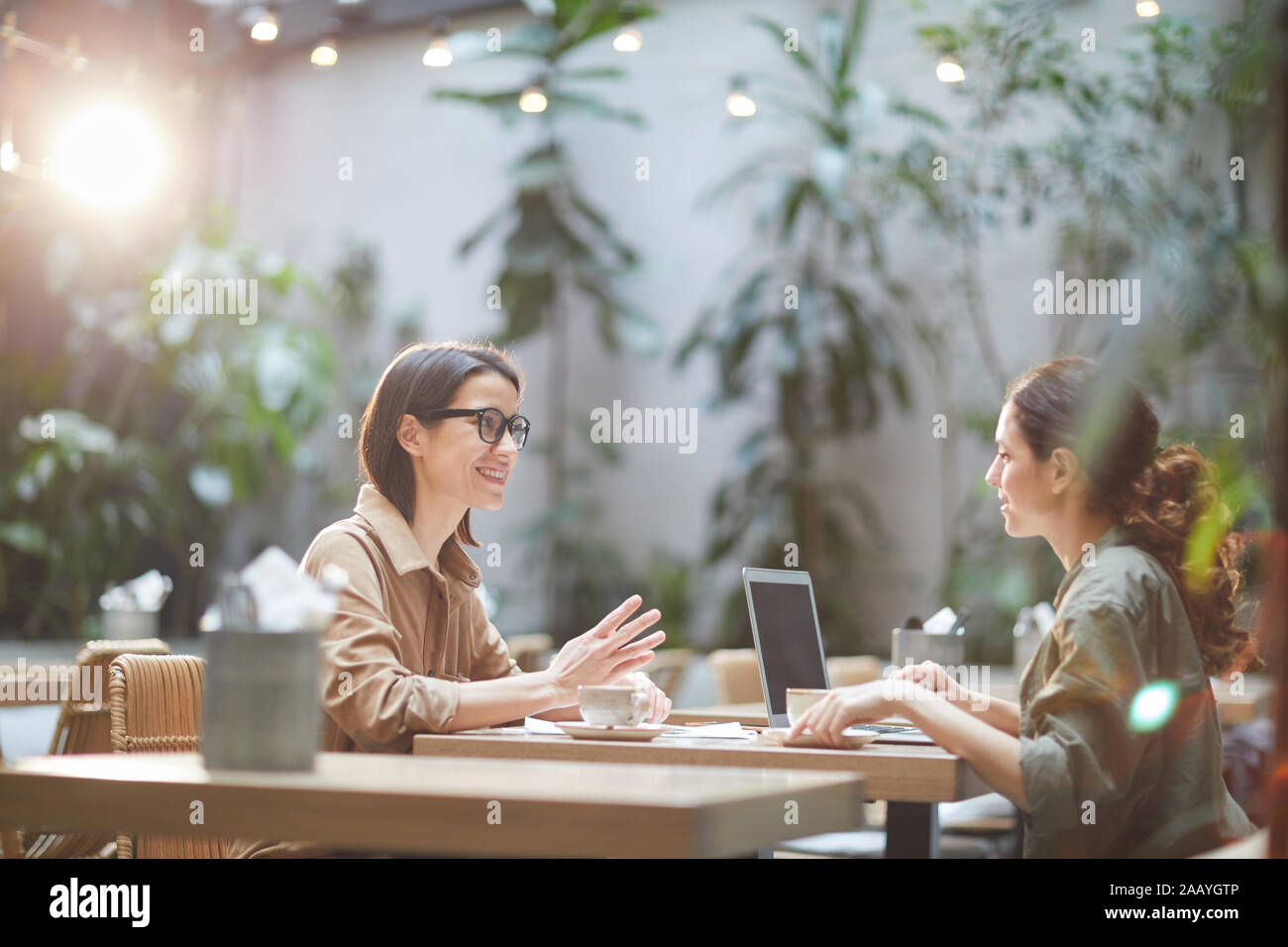 Side view portrait of two modern young women sitting at table in cafe and smiling cheerfully enjoying lunch together, copy space Stock Photo