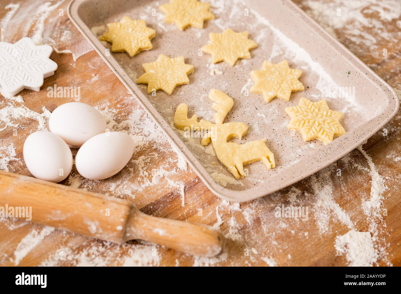 Tray with two rows of raw Christmas cookies with three eggs, rolling pin, flour and cutters near by on table Stock Photo