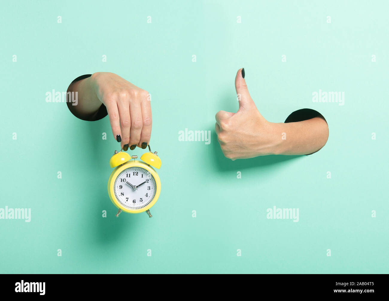 Female hand hold alarm clock and show tumb sign through a hole on neon mint background. Just in time concept. Stock Photo