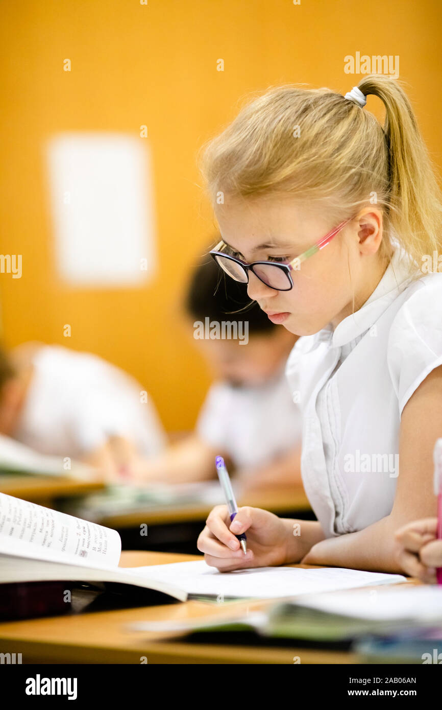 Smiling scholar girl sitting with other children in classroom and writing on textbook. Happy student doing homework at elementary school. Young school Stock Photo