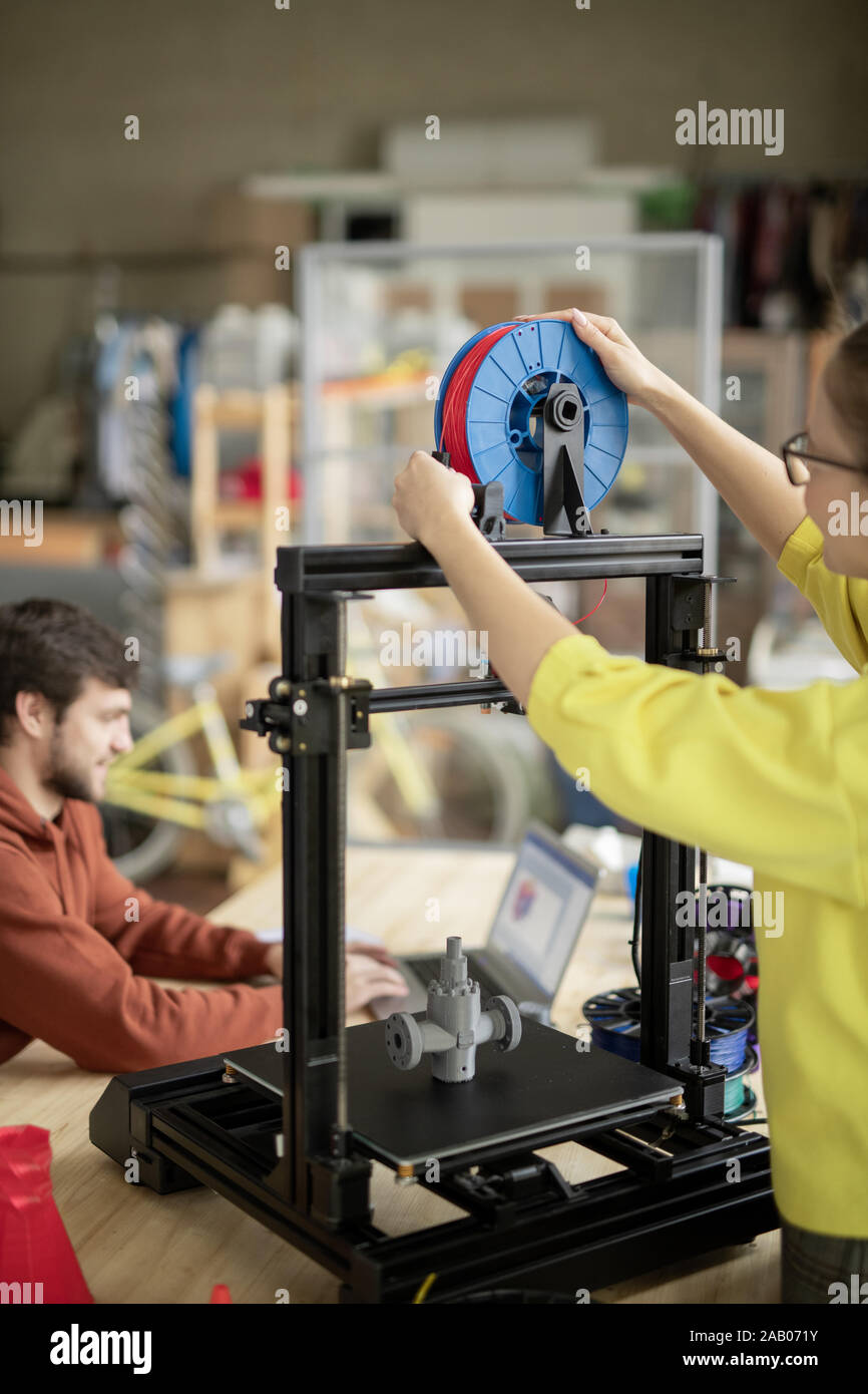 Young woman putting new spool with red filament on 3d printer while her colleague searching for online ideas on background Stock Photo