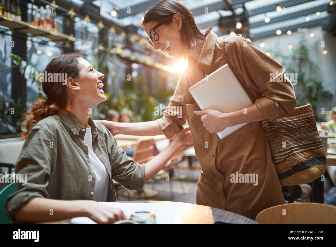 Side view portrait of two young women greeting each other joyfully during meeting in cafe on outdoor terrace decorated with lights Stock Photo